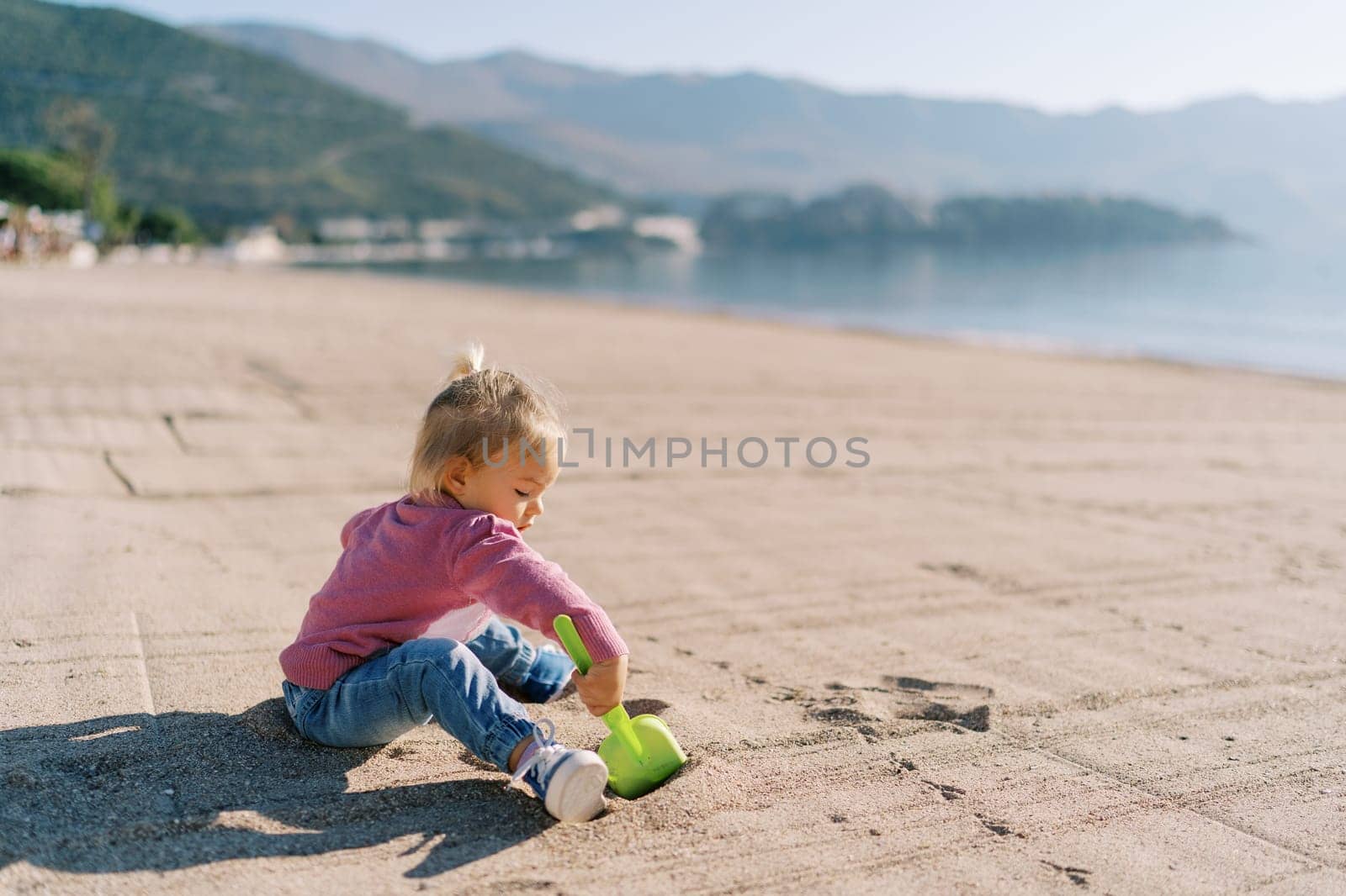 Little girl sits on a sandy beach and digs the sand with a toy shovel. High quality photo