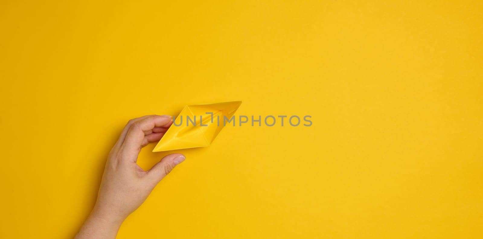 Female hand hold a blue paper boat on a yellow background. Mentoring and support concept by ndanko