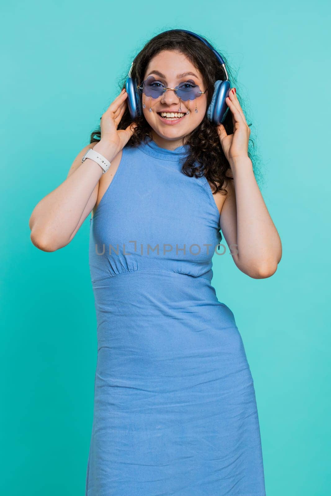 Happy woman in headphones listening music and dancing disco fooling around having fun expressive gesticulating hands relaxing on party. Girl in sunglasses isolated on studio blue background. Vertical