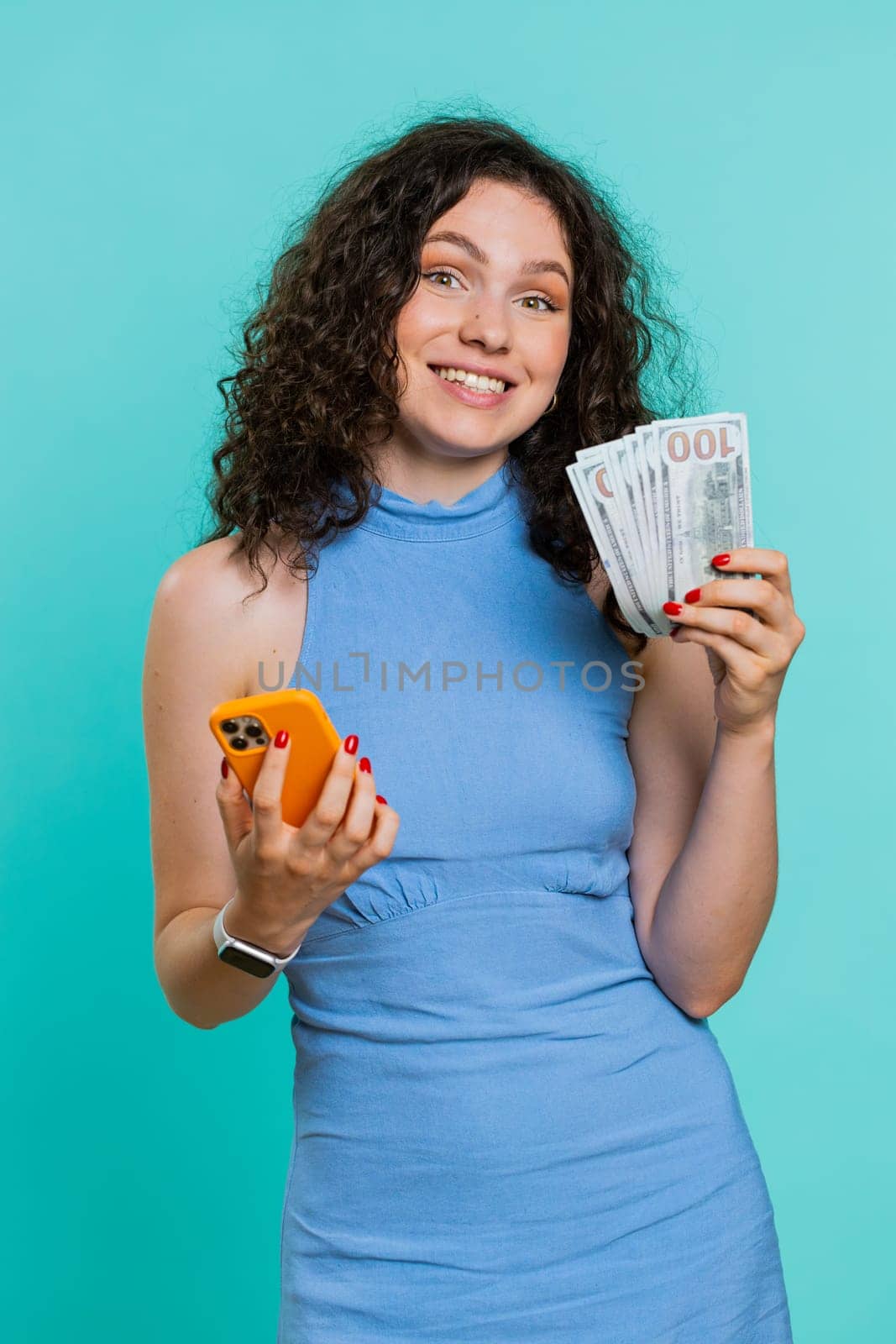 Young woman looking smartphone display sincerely rejoicing win, receiving money dollar cash banknotes, celebrating success lottery luck. Adult girl isolated alone on blue studio background. Vertical
