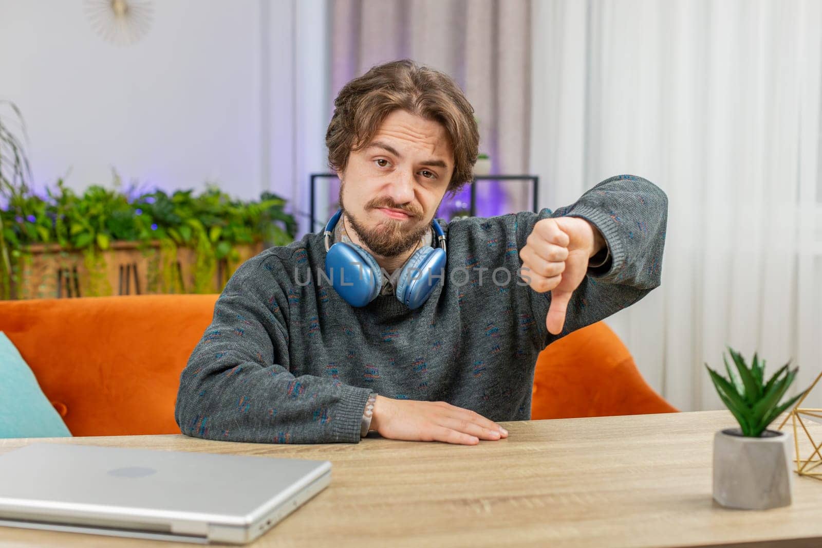 Dislike. Upset unhappy Caucasian man showing thumbs down sign fail gesture, expressing discontent, disapproval dissatisfied bad work at home office indoors. Displeased young guy in room sits at table