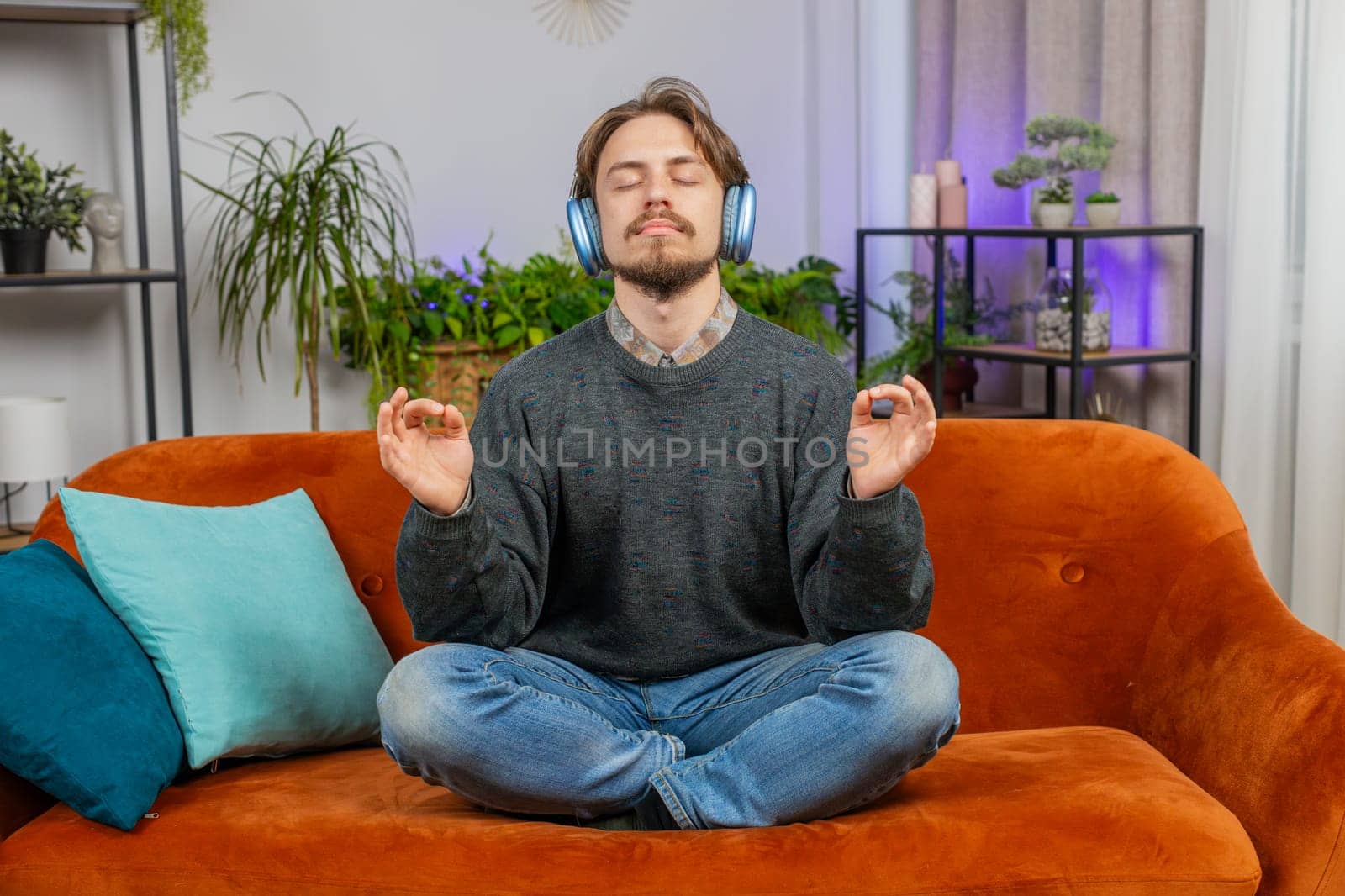 Keep calm down, relax. Bearded young man listening music breathes deeply, eyes closed meditating with concentrated thoughts, peaceful mind. Young Caucasian guy sits at home couch in room apartment