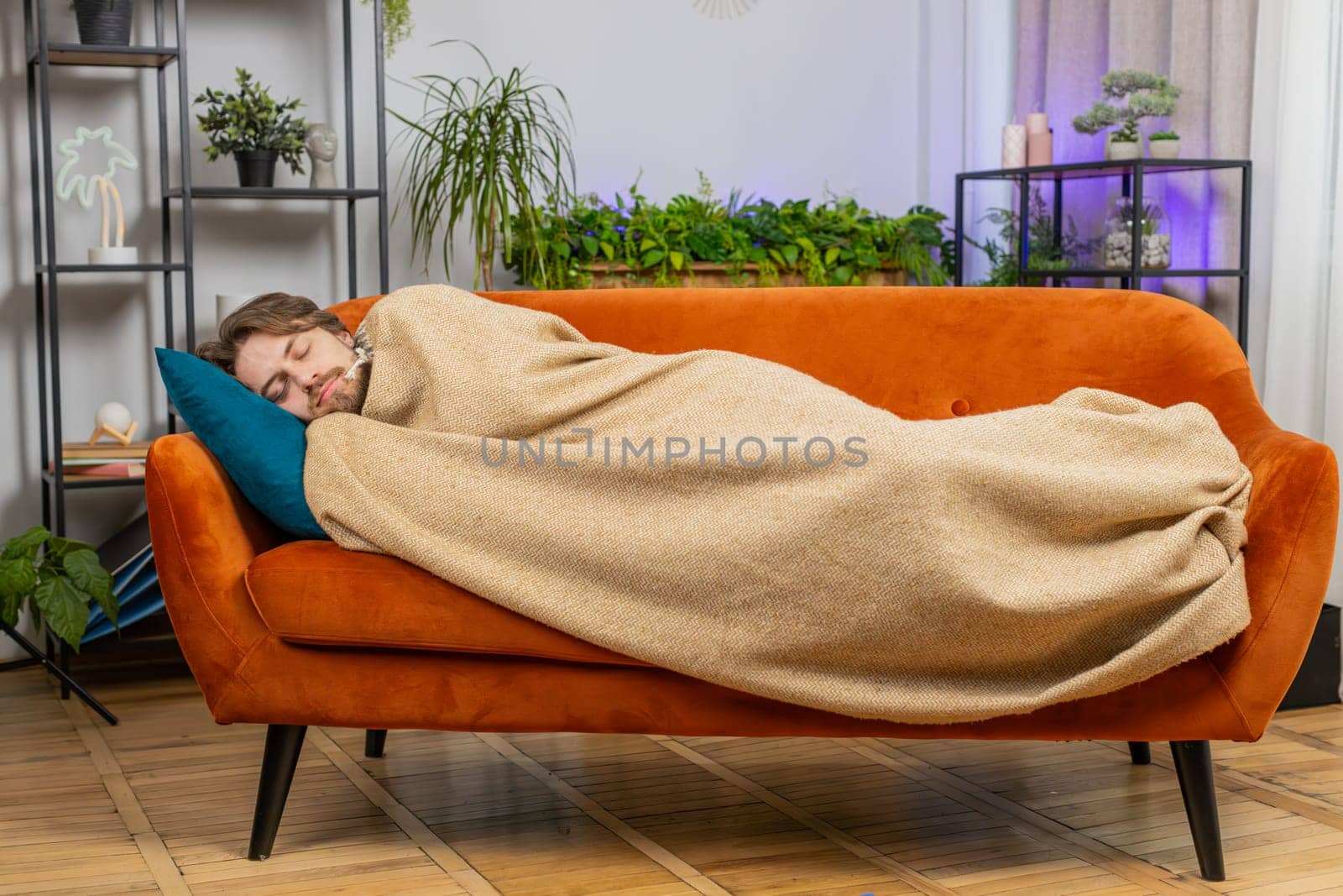 Tired young man lying down in bed taking a rest at home apartment room. Carefree Caucasian guy napping, falling asleep on comfortable orange sofa with pillows. Closed his eyes enjoy daytime nap alone