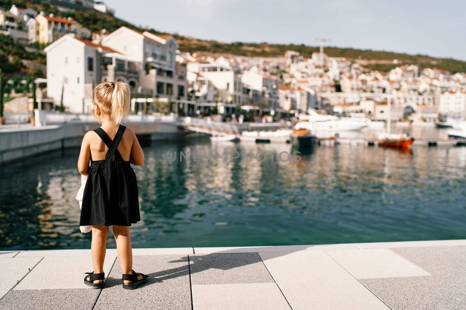 Little girl stands on the pier and looks at the marina with yachts. Back view. High quality photo