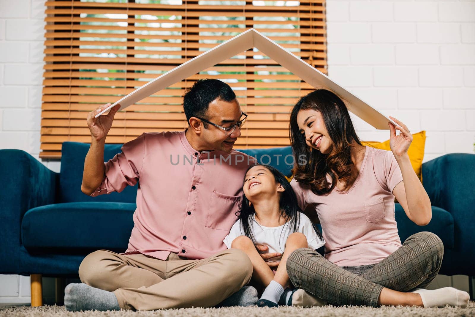 In their new house a loving family holds a cardboard roof on a couch symbolizing investment and happiness. Asian parents and their daughter radiate joy and security.