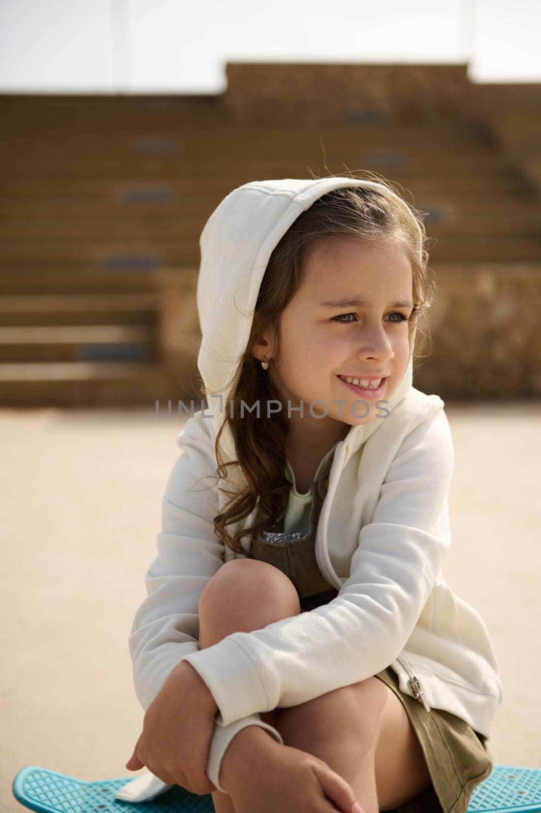 Close-up stylish little kid girl sitting on her blue skateboard, smiling dreamily looking away, dressed in beige hoodie by artgf