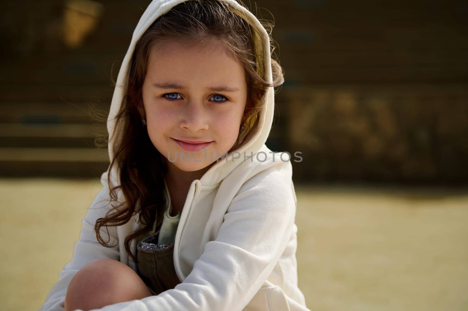 Close-up portrait adorable little child girl with blue eyes, wearing beige hoodie with a hood on her head, smiling looking confidently at camera, sitting on a skateboard in outdoor urban skatepark