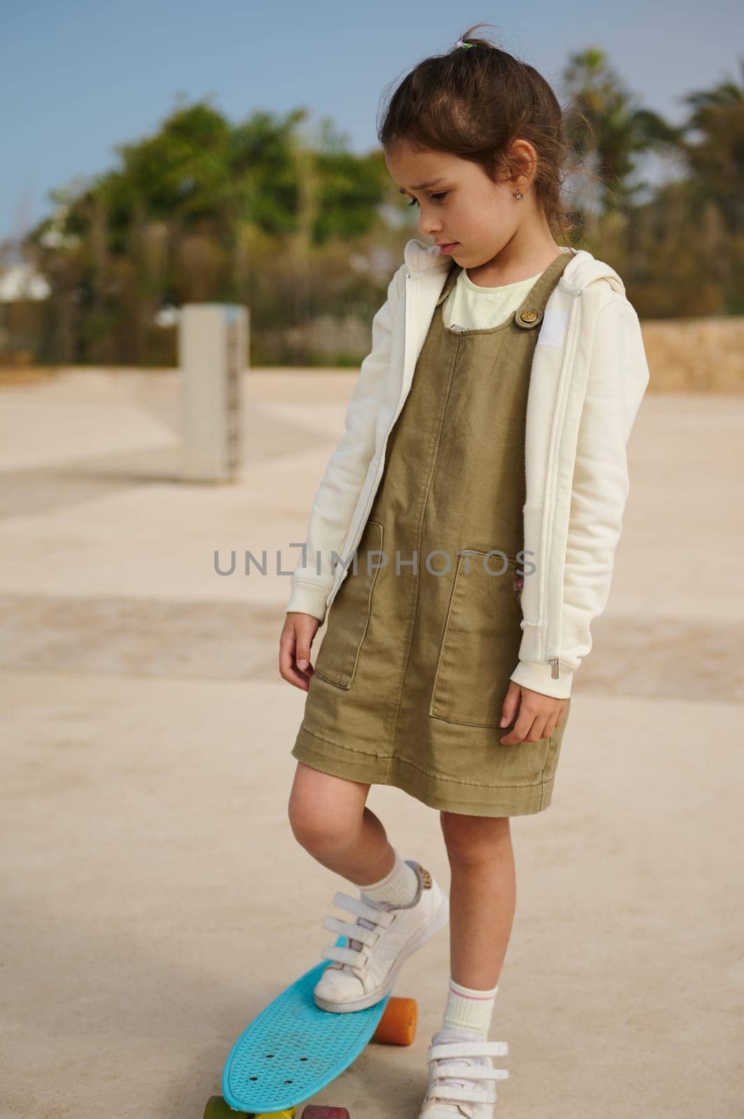 Beautiful little child girl standing on her skateboard on one leg, expressing sad emotions, feeling upset while having difficulties on learning skateboarding, standing alone on a skatepark playground