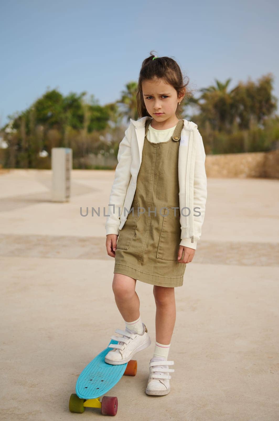 Upset Caucasian child girl standing on her skateboard on one leg, expressing sad emotions, looking at camera, feeling upset having difficulties on skateboarding, standing alone on skatepark playground