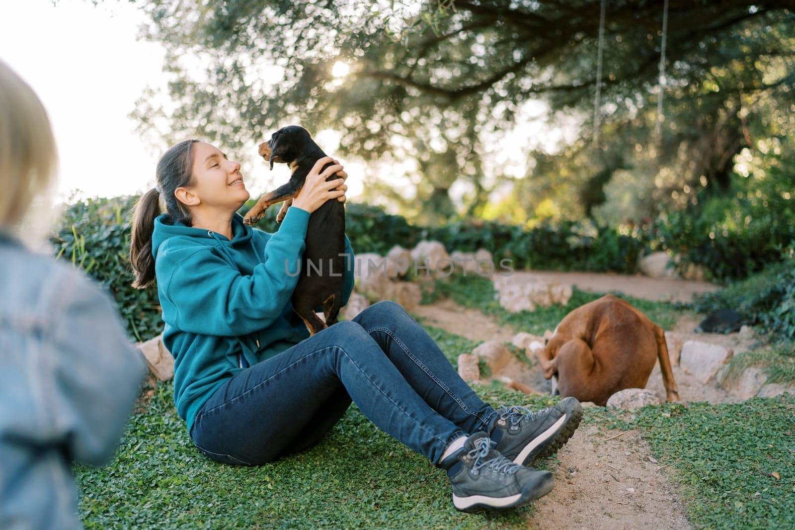Little girl looks at her mother raising a black puppy in her arms while sitting on the grass. High quality photo