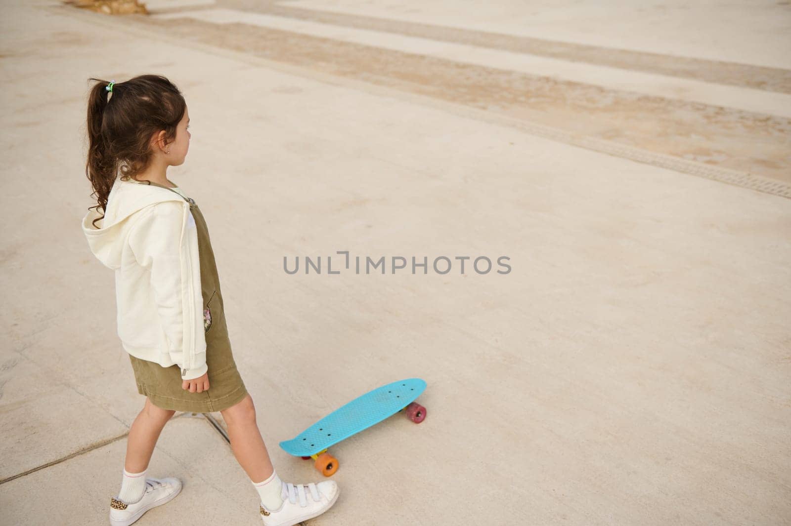 Little child girl in beige hoodie and khaki dress, skateboarding on the urban skatepark or playground. View from above. Happy active childhood. Extreme sport. Leisure activity