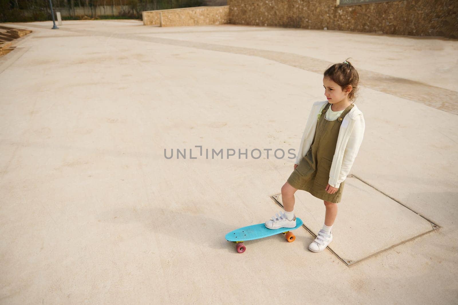 View from above of a beautiful little girl standing on her skateboard on one leg, dreamily looking into the distance by artgf