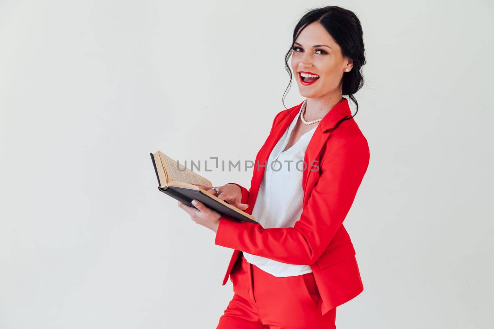 A young woman in a business suit, with a red book in hand, sitting on the wooden floor with a thoughtful face, glance to the side, full height.