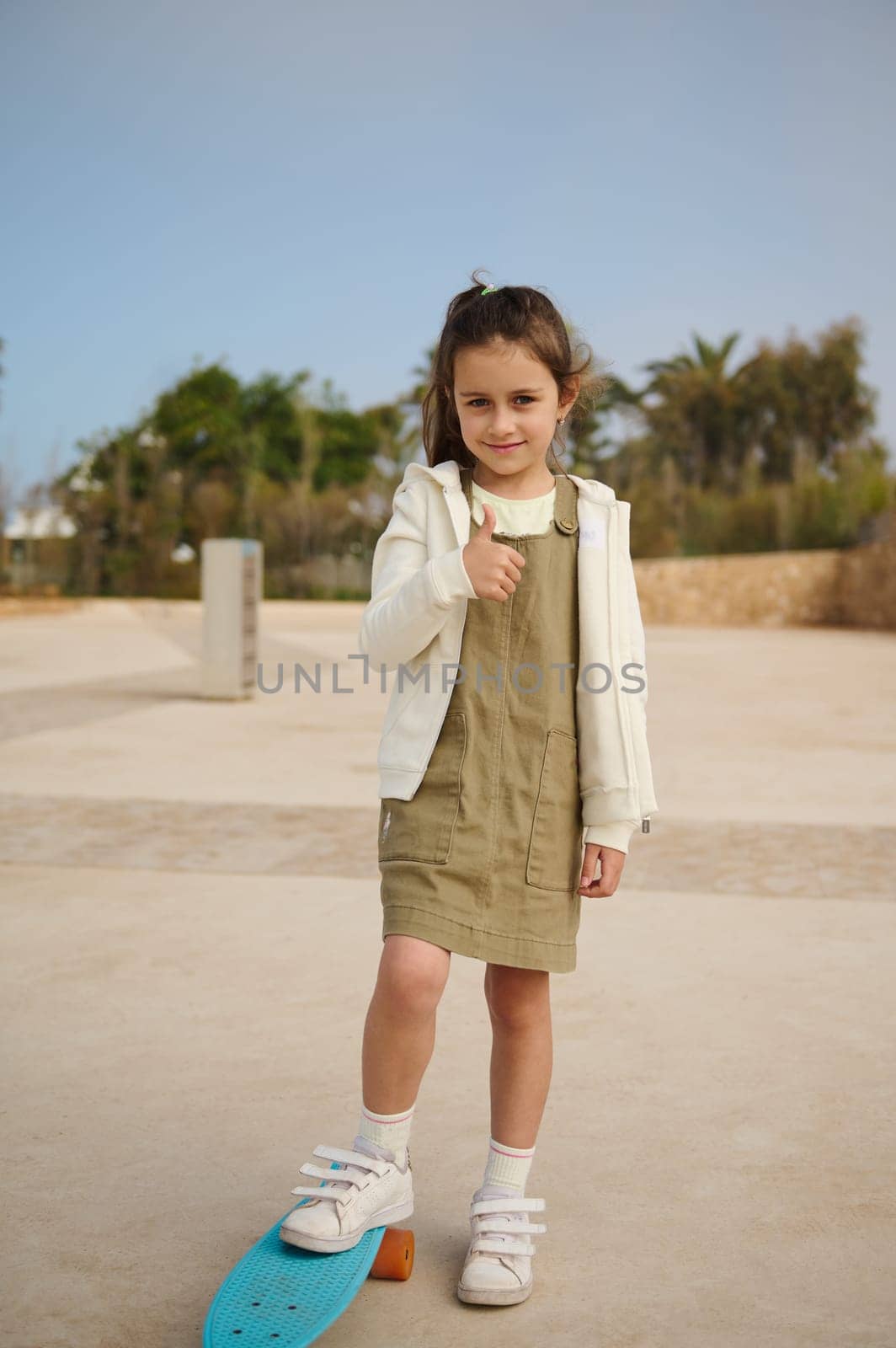 Happy Caucasian elementary age child girl smiles cutely, looking at camera, gestures with hand sign, shows a thumb up, expressing positive emotions, standing on her skateboard on one leg at skatepark