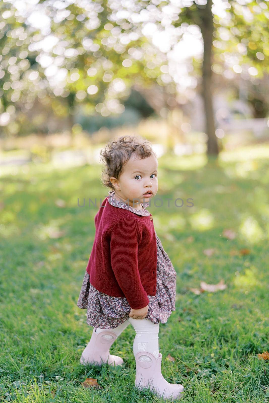 Little girl is standing on the green grass in the garden, looking interestedly to the side by Nadtochiy