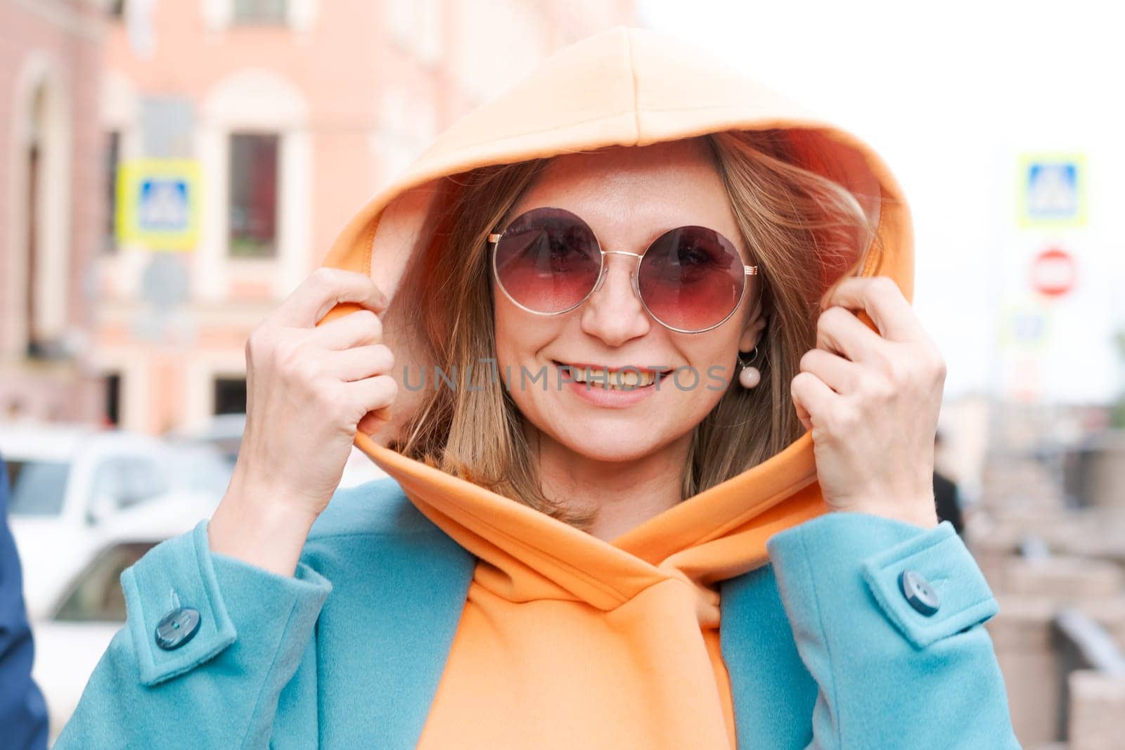 Happy woman walking through city with bag in her hand, dressed in bright clothes, an orange hoodie and a blue coat, smiling contentedly in sunglasses