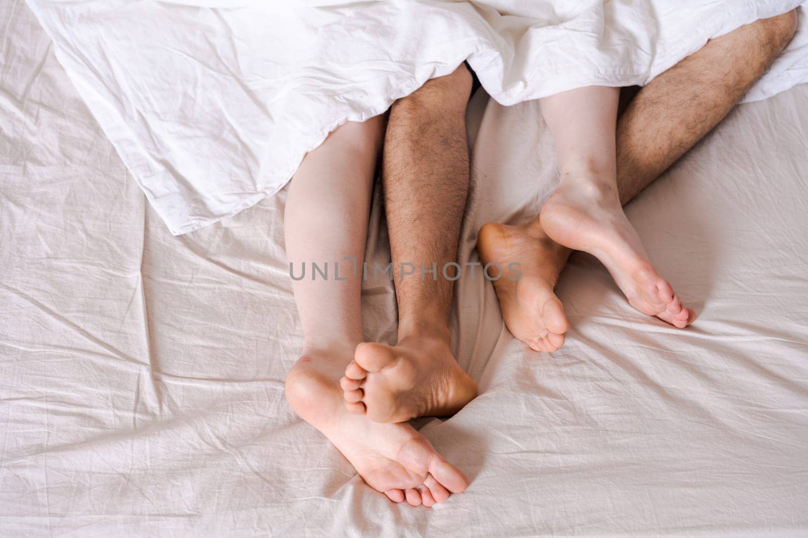 Feet of a couple in bed under the white blanket. by EkaterinaPereslavtseva