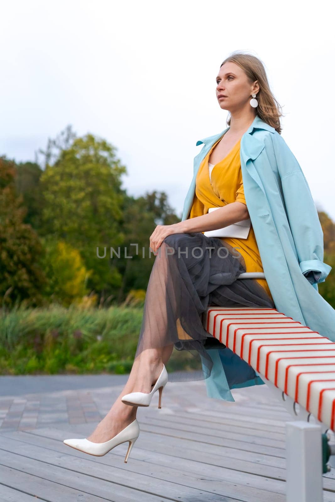Happy woman swinging on swing in city park, wearing yellow dress and blue coat by EkaterinaPereslavtseva