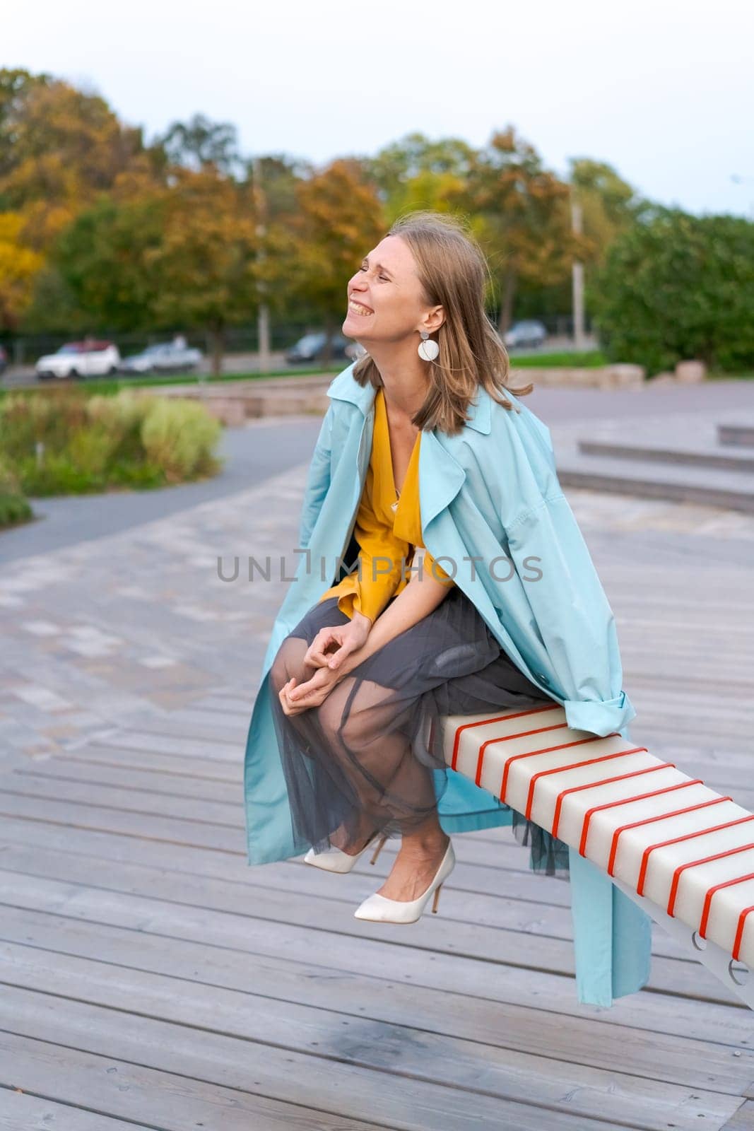 Happy woman swinging on swing in city park, wearing yellow dress and blue coat, the concept of freedom from worries and problems
