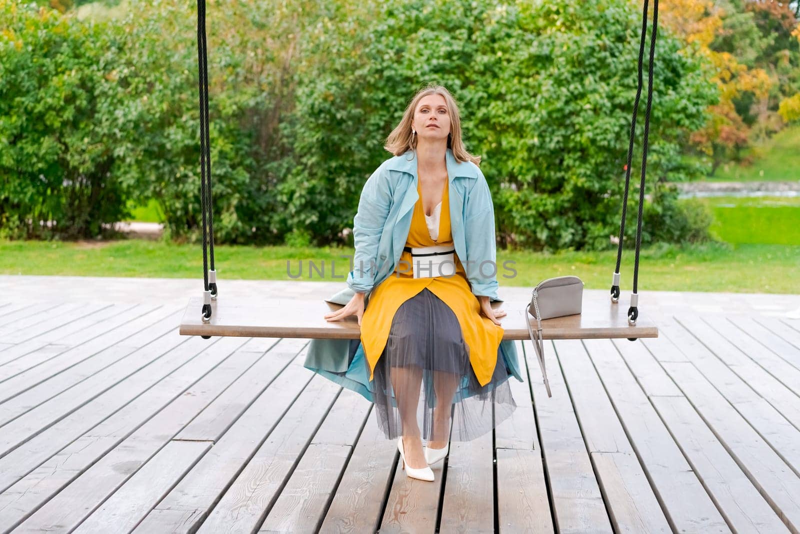 Happy woman swinging on swing in city park, wearing yellow dress and blue coat by EkaterinaPereslavtseva