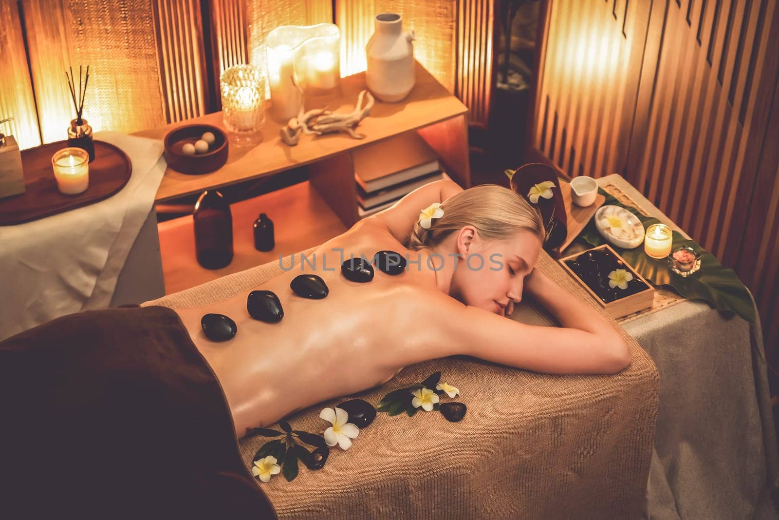 Hot stone massage at spa salon in luxury resort with warm candle light, blissful woman customer enjoying spa basalt stone massage glide over body with soothing warmth. Quiescent