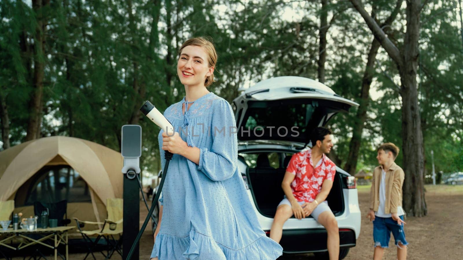 Outdoor adventure and family vacation camping at sea travel by eco friendly car. Cheerful woman or mother holding, pointing EV charger point with playful and happiness posture in campsite. Perpetual