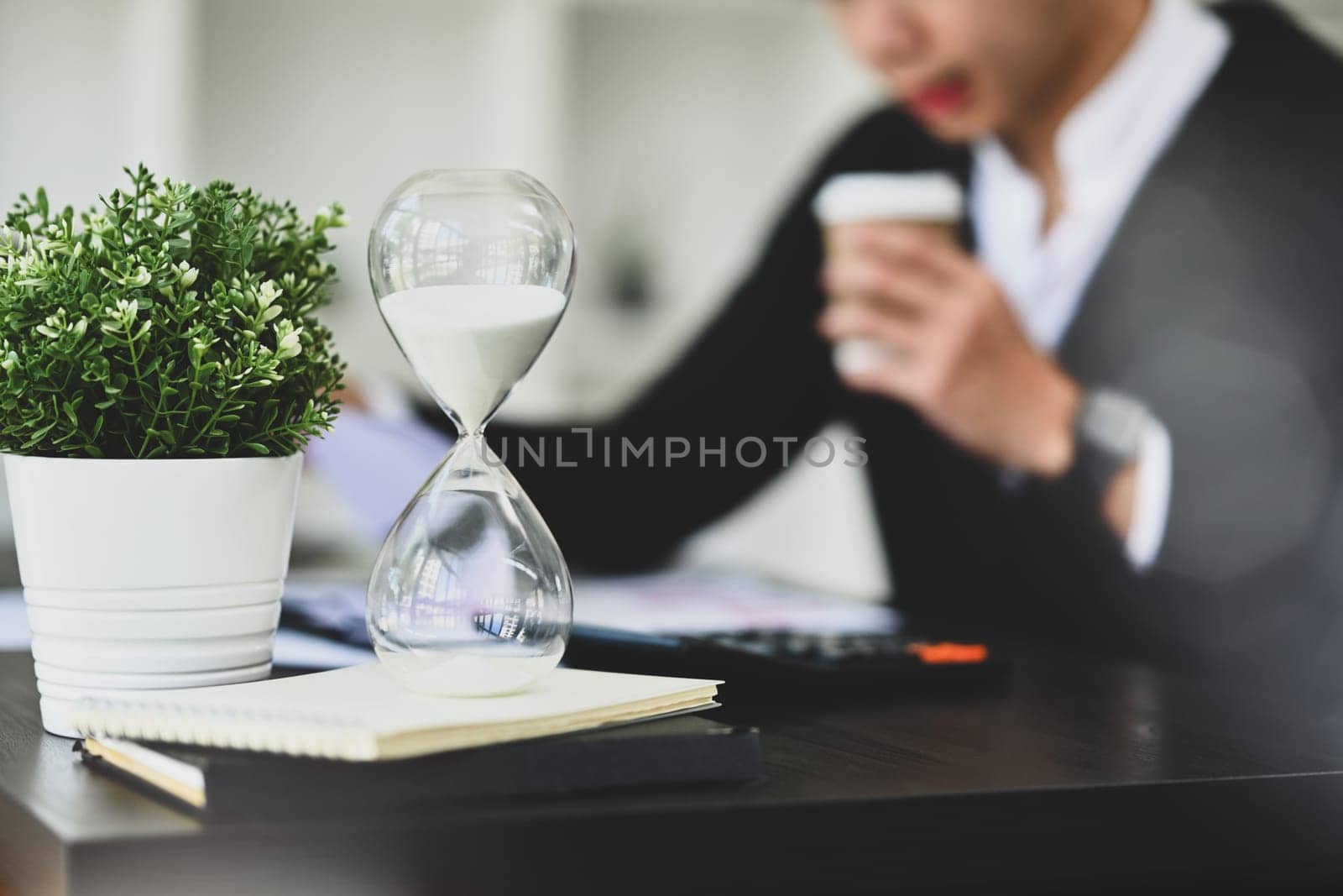 Hourglass on black office table with blurred businessman working on background. Time management concept by prathanchorruangsak