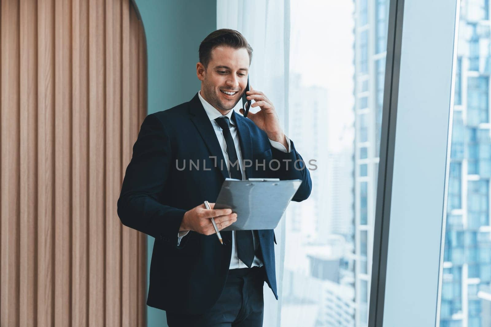 Portrait of professional businessman stand near window with skyscraper while phone calling and holding laptop. Busy executive manager holding laptop while multitasking working. Closeup. Ornamented.