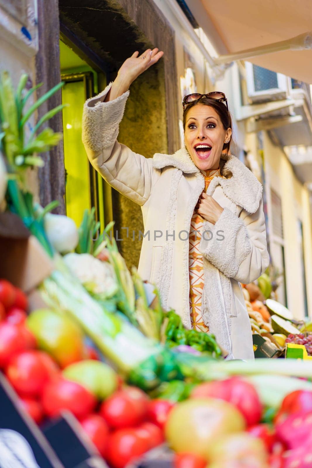 Low angle of positive young woman in warm clothes, smiling and looking at camera while waving hand and standing near stall with various vegetables and fruits in daylight