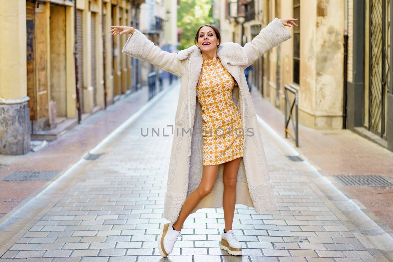 Stylish young trendy woman in long coat and dress dancing on street by javiindy