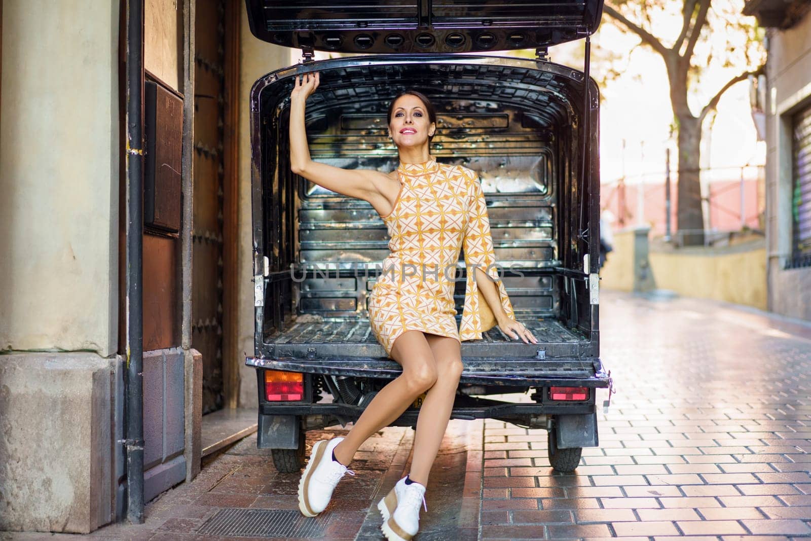 Full body of confident young woman in stylish short dress and sneakers, sitting on empty metallic back of van on paved street in daylight while raising hand and looking at camera