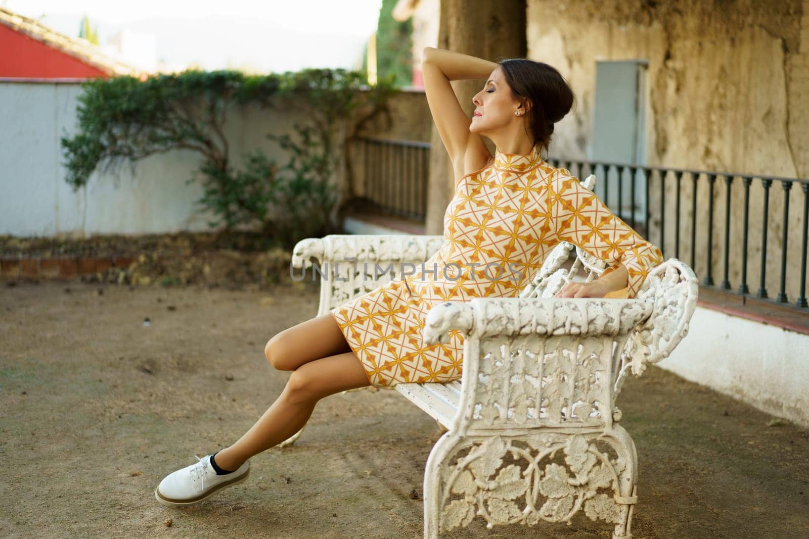 Full body of young alluring female model in stylish dress and sneakers sitting up in romantic pose with closed eyes on bench by old building