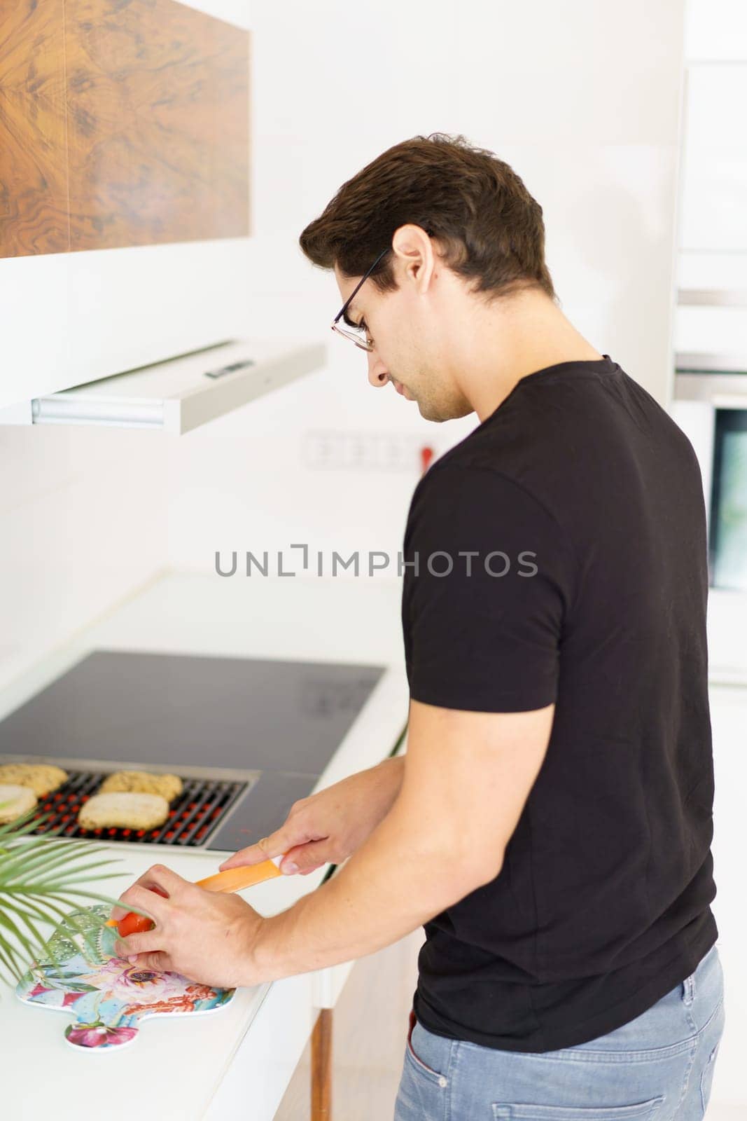 Side view of adult male in casual clothes and eyeglasses looking down while standing in kitchen near cooking range, with bread slices on grill and cutting tomato with knife in daylight
