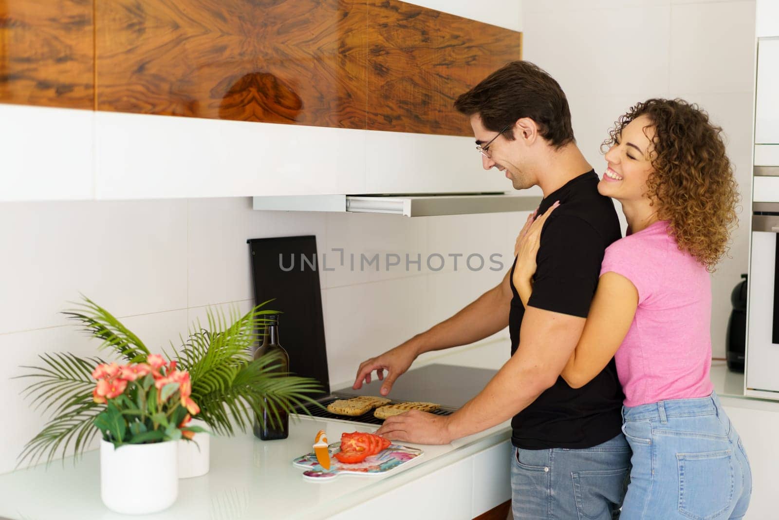 Side view of happy adult couple while standing near cooking range smiling looking down male in eyeglasses toasting bread slices, on grill and female with eyes closed hugging from behind in daylight