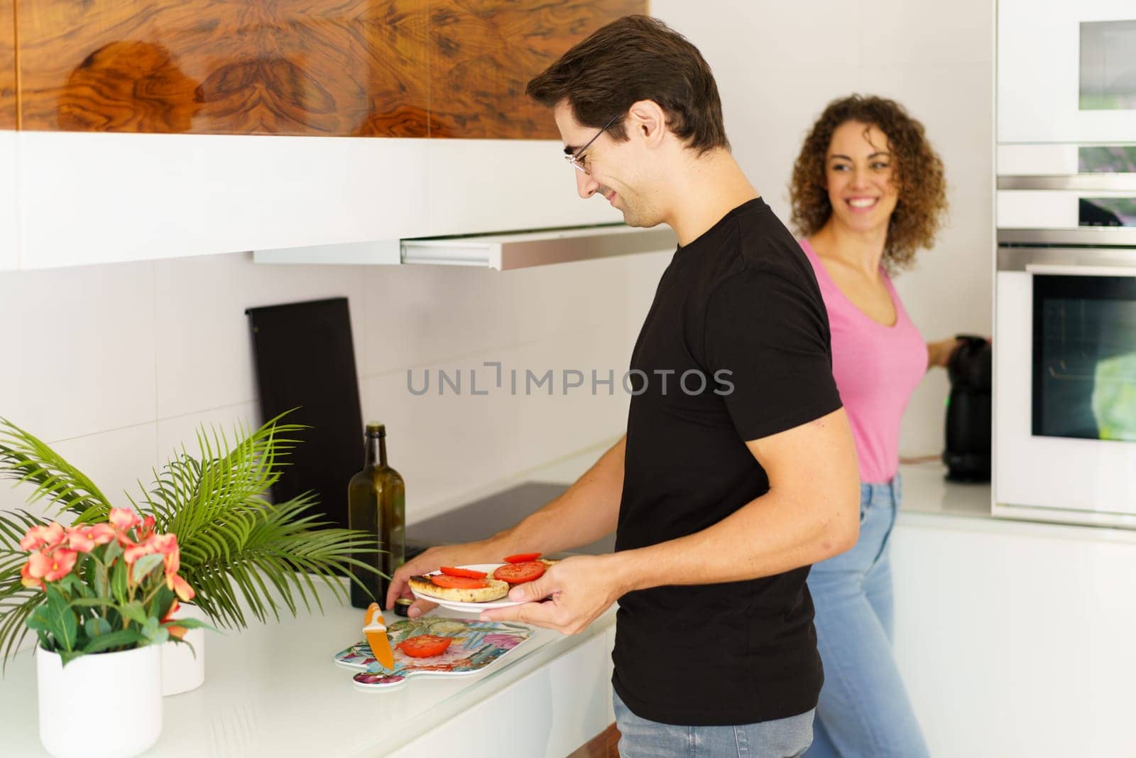 Side view of happy adult male looking down while standing with smiling conversing female at counter, with wine bottle flower pot and preparing healthy salad in kitchen at home in daylight