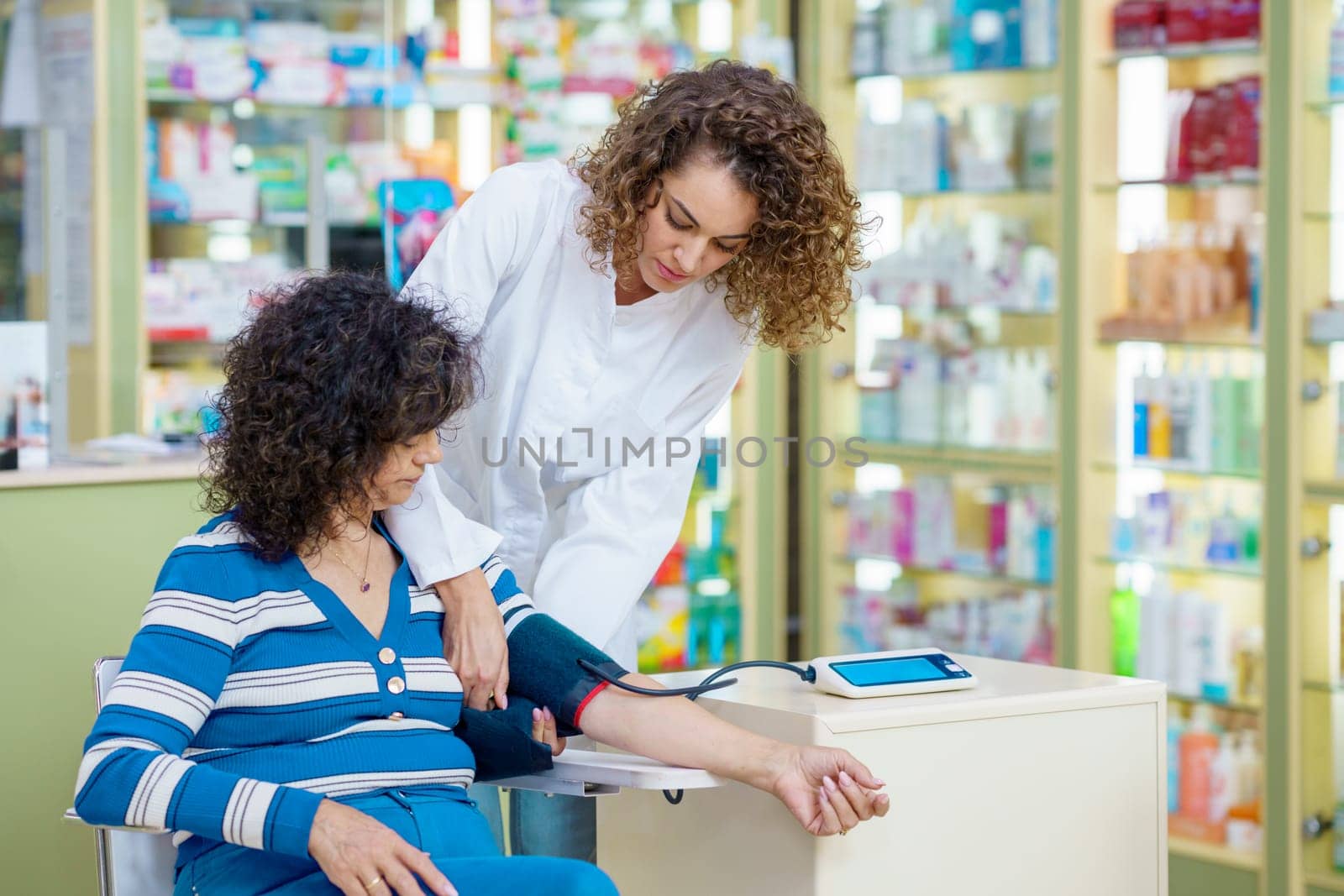 Young woman pharmacist checking blood pressure of lady customer in pharmacy by javiindy