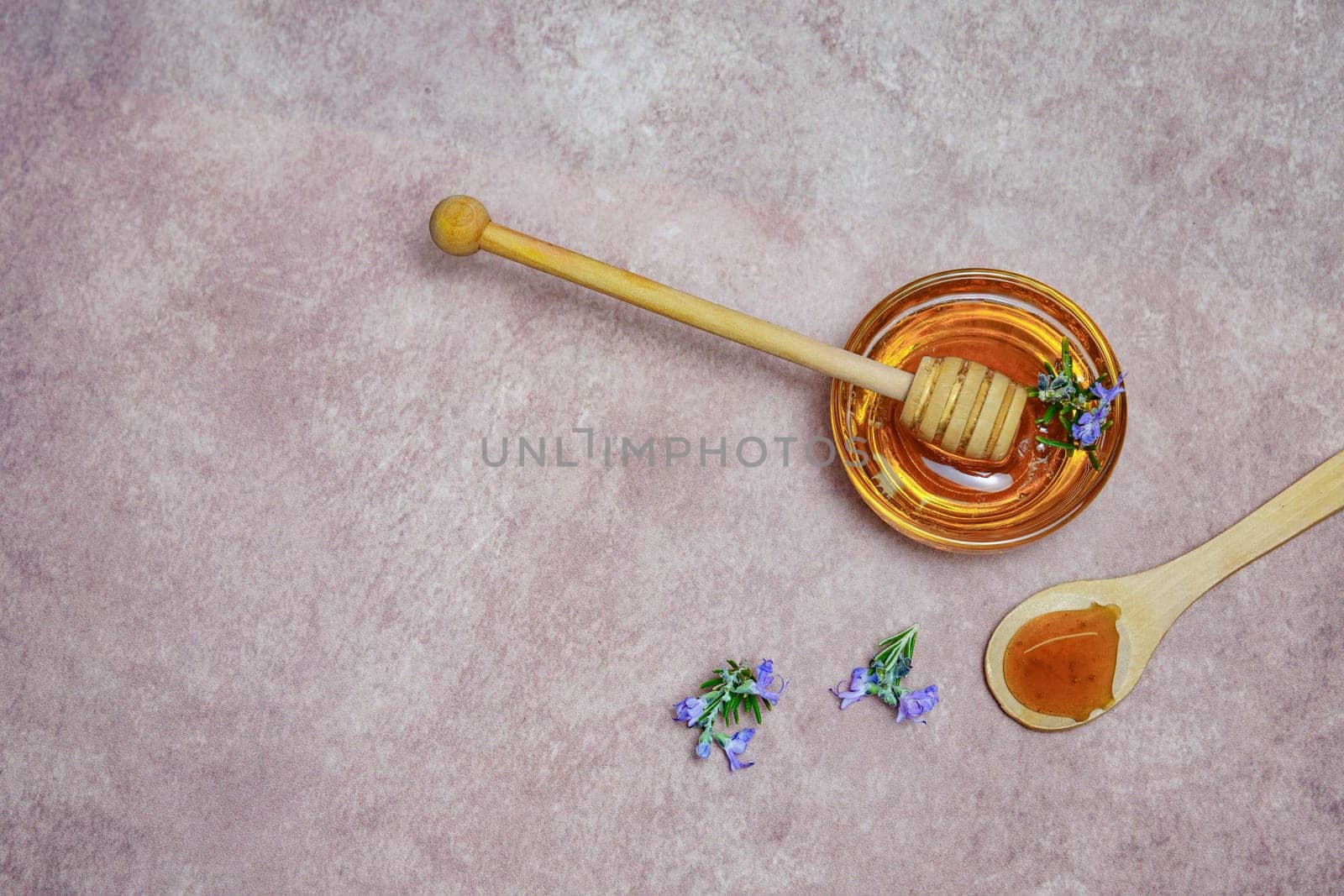 top view of natural rosemary honey in a glass bowl with a wooden spoon and fresh rosemary branches in blossom