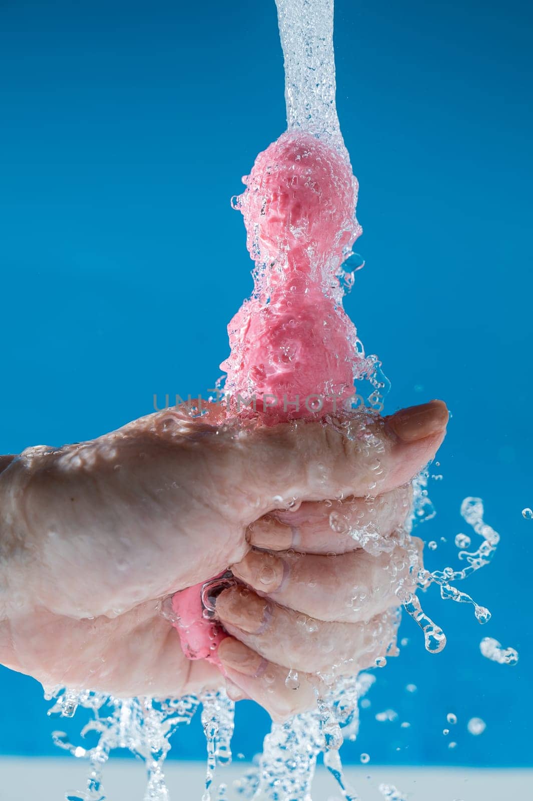 Woman holding pink anal beads under running water on blue background. Sex toy hygiene concept