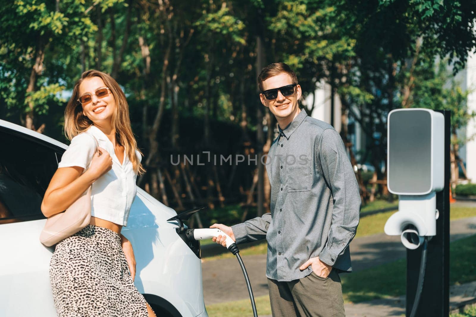 Young couple travel with EV electric car charging in green sustainable city outdoor garden in summer shows urban sustainability lifestyle by green clean rechargeable energy of electric vehicle innards