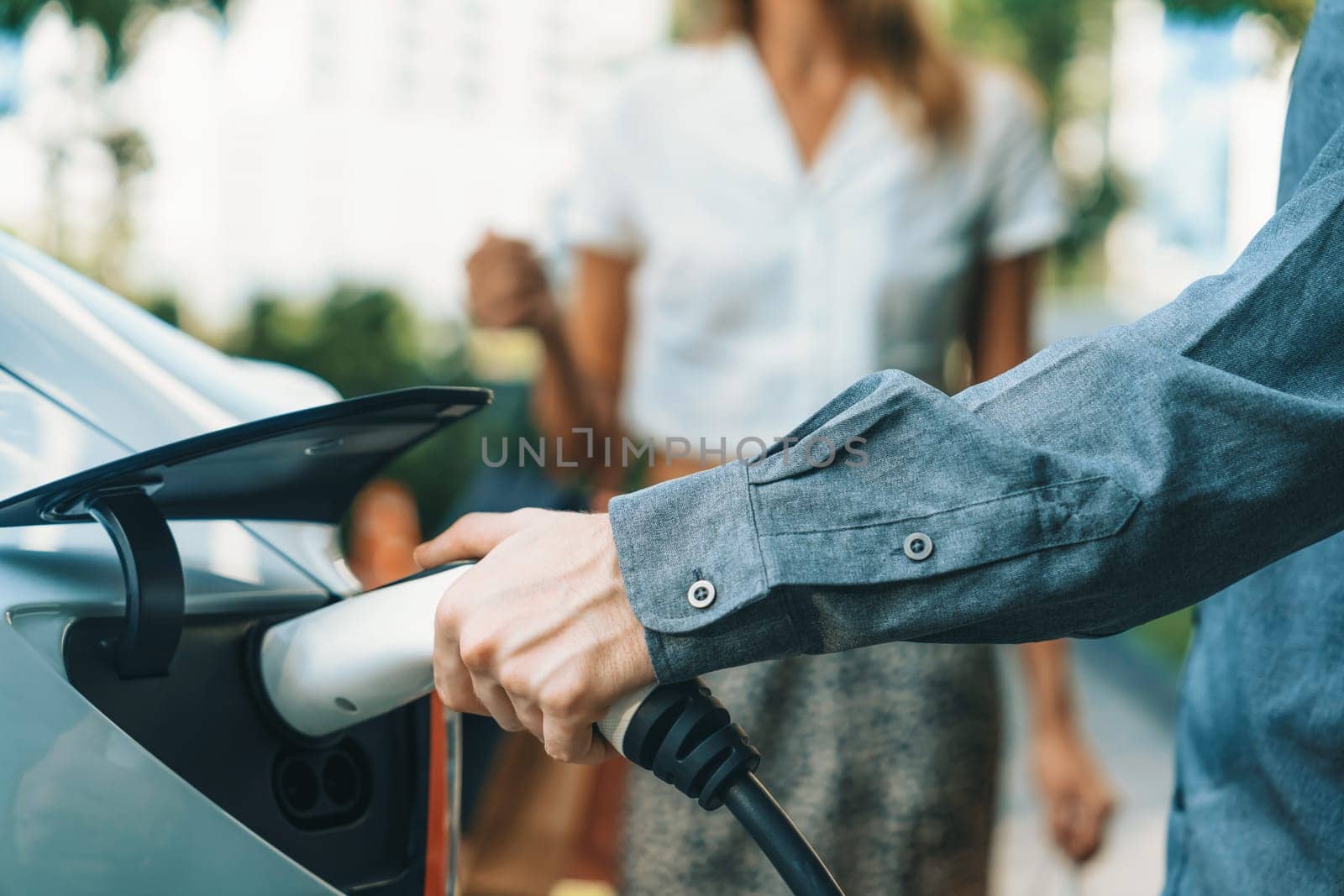 Young couple travel with EV electric car charging in green sustainable city outdoor garden in summer shows urban sustainability lifestyle by green clean rechargeable energy of electric vehicle innards