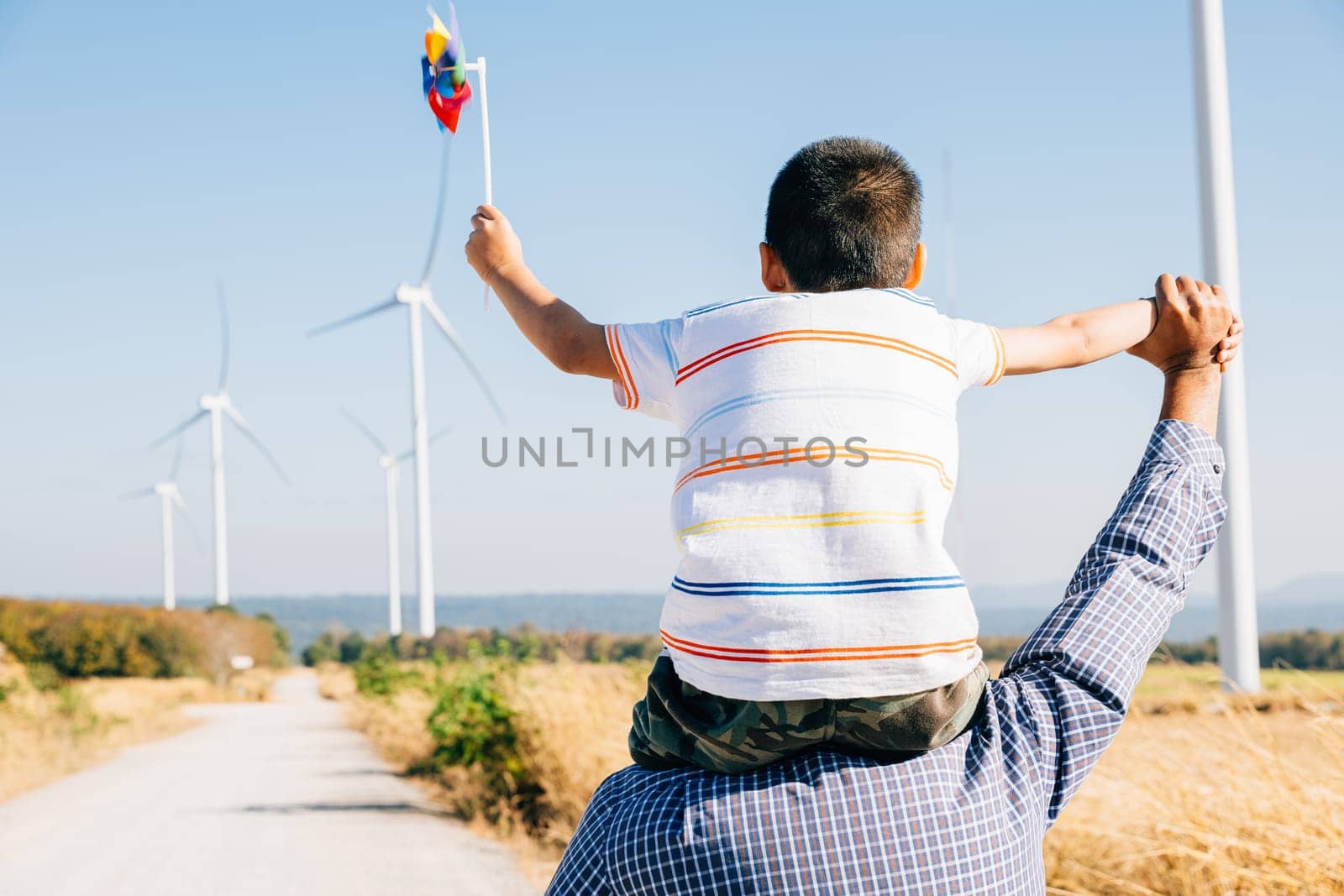 Father smiles carrying daughter with pinwheel. Family joy near windmills embodies global innovation in renewable energy. A bonding moment filled with happiness and community. Father Day