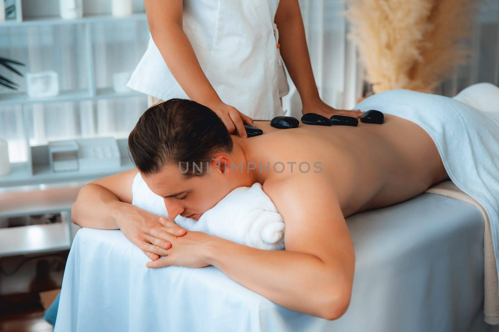 Hot stone massage at spa salon in luxury resort with day light serenity ambient, blissful man customer enjoying spa basalt stone massage glide over body with soothing warmth. Quiescent