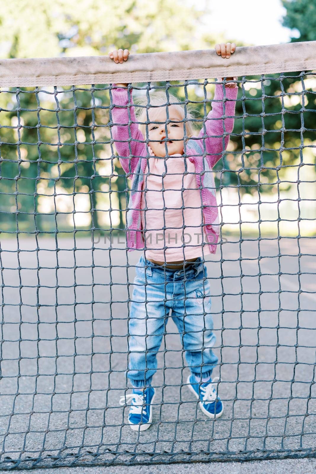 Little girl clings to the edge of the tennis net with her hands, standing on tiptoe. High quality photo