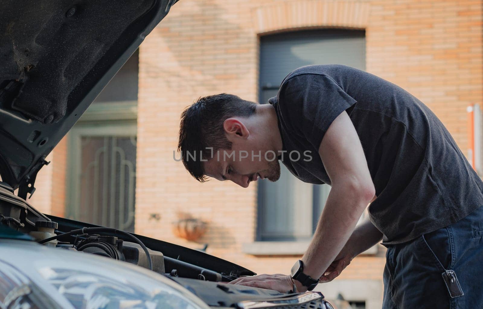 One young caucasian guy looks into the open hood of his car while standing on a city street on a summer afternoon in the evening, close-up side view. The concept of replacing light bulbs, car repair.