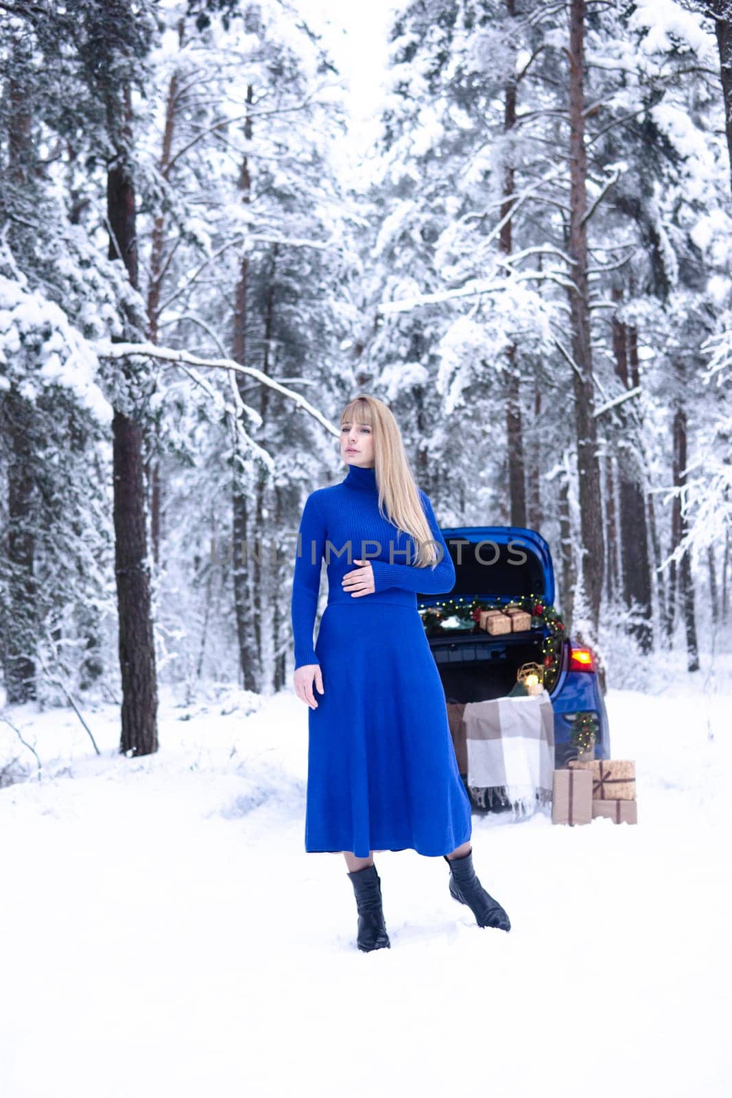 Woman in winter snowy forest in blue dress next to blue car decorated with Christmas decor. Christmas and winter holidays concept