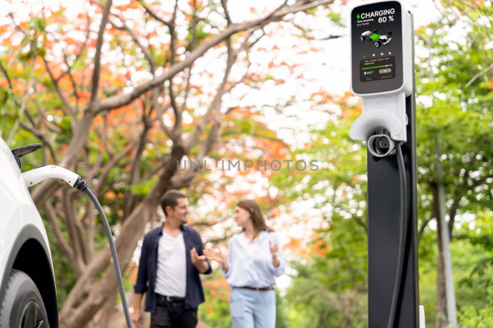 Focused EV car recharging battery on blurred background of lovey couple during autumnal road trip travel with electric vehicle recharging battery. Eco friendly travel on vacation during autumn. Exalt