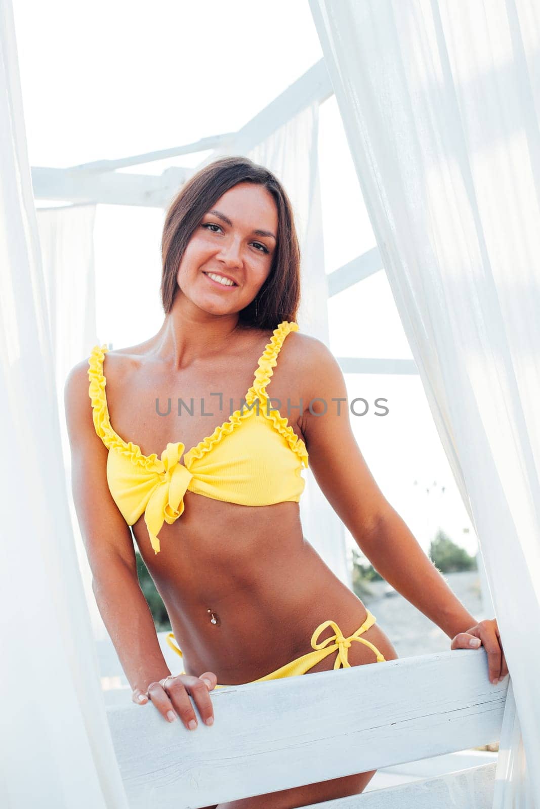 Tanned woman in yellow swimsuit with long hair sits in the shade on the beach by Simakov