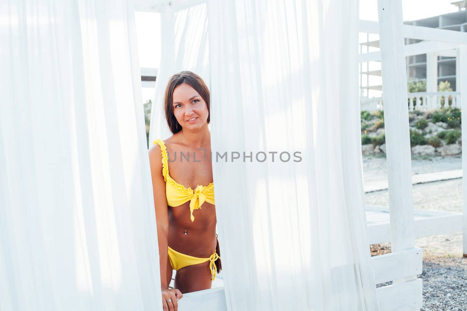 Tanned woman in yellow swimsuit with long hair sits in the shade on the beach by Simakov