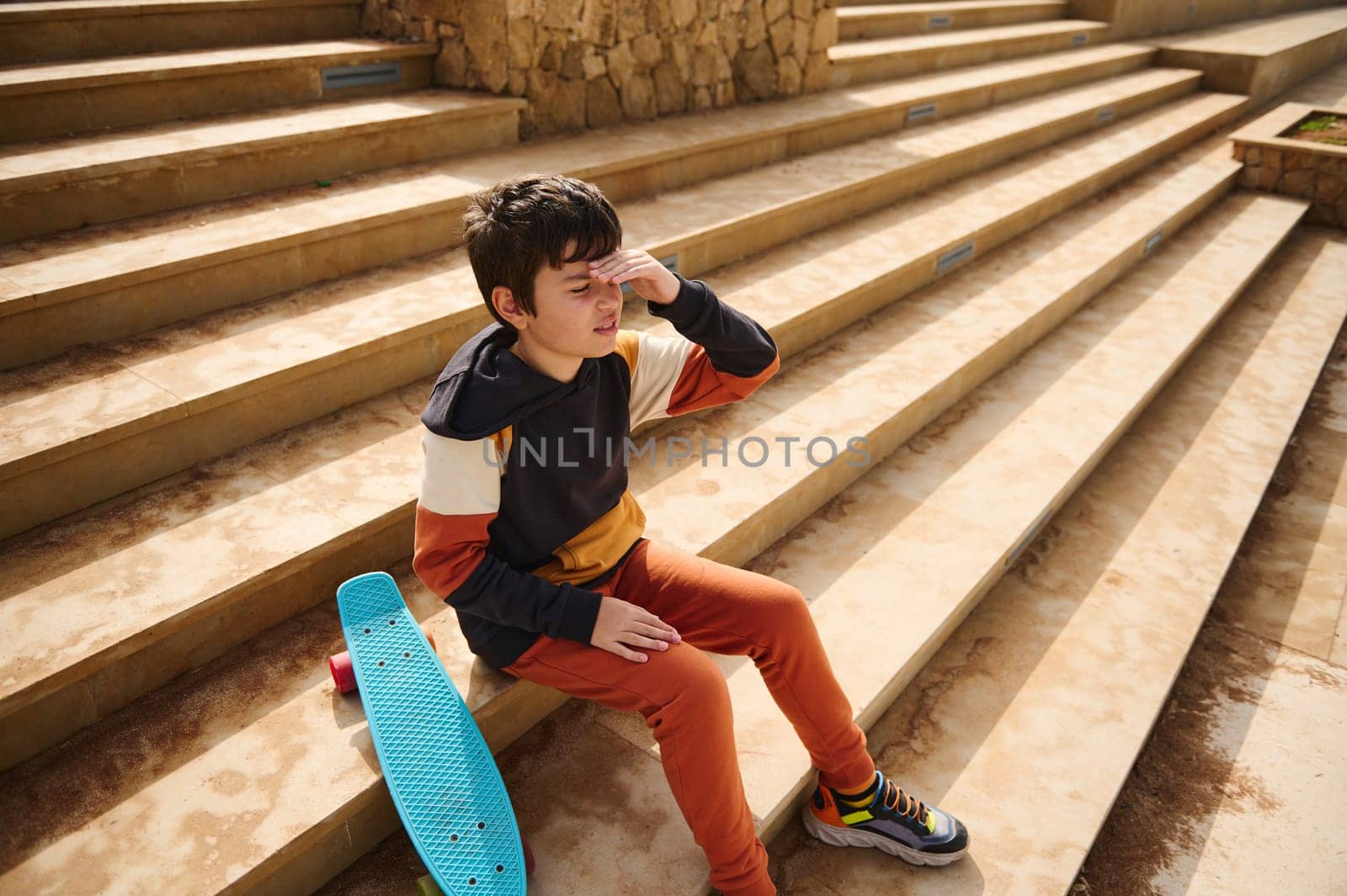 Teenage student boy, schoolboy in sportswear sitting on steps in the urban skatepark, thoughtfully looking into distance by artgf