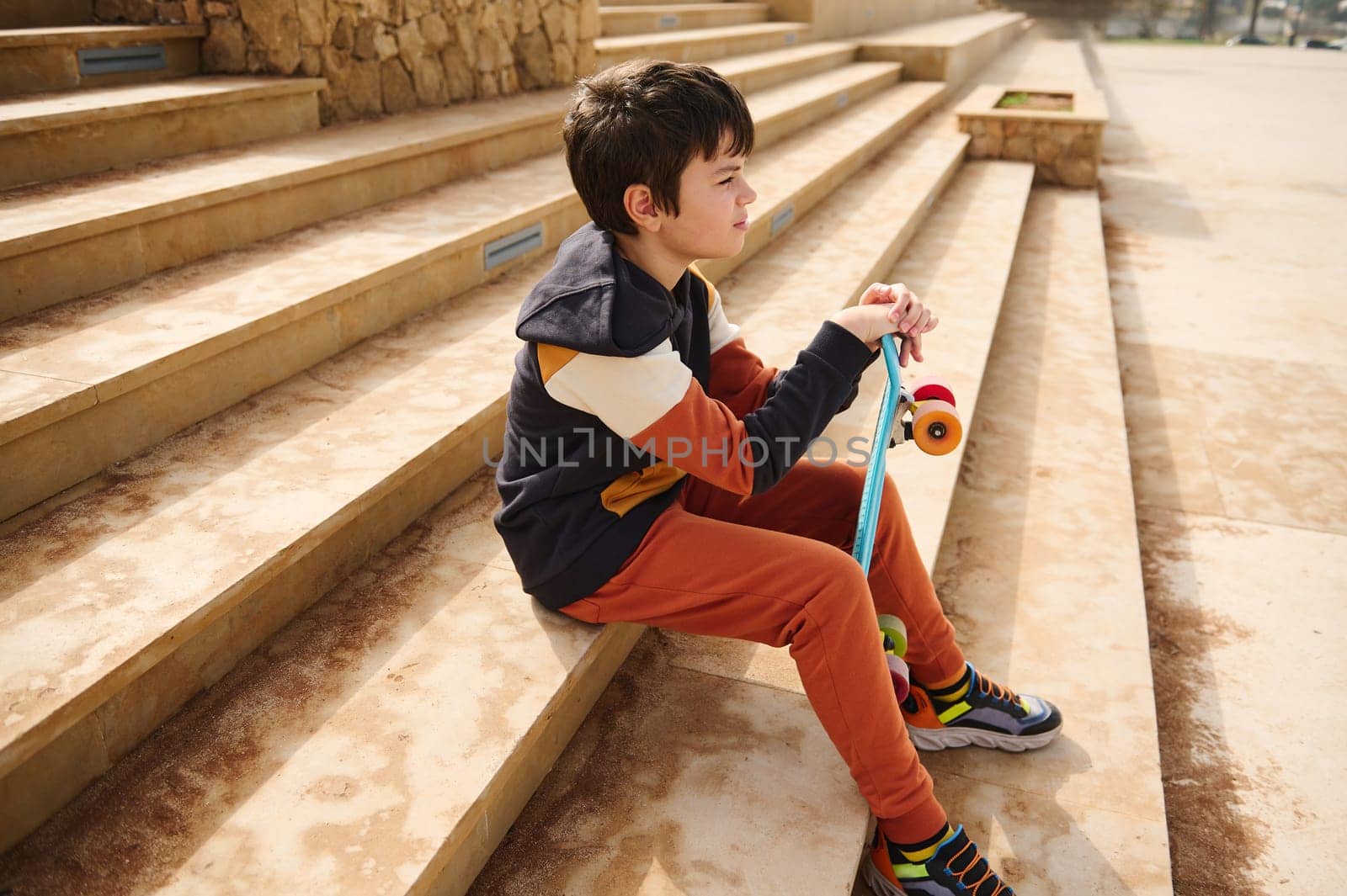 Teenage student boy, schoolboy in sportswear sitting on steps in urban skatepark, thoughtfully looking into the distance by artgf